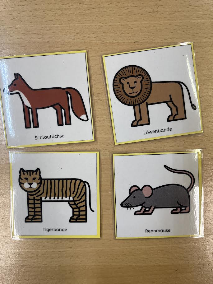 Das Foto zeigt vier quadratische Kärtchen mit bunten Tierzeichnungen, die auf einem Holztisch liegen. Oben links ist ein roter Fuchs mit buschigem Schwanz zu sehen, darunter steht „Schlaufüchse“. Oben rechts ist ein brauner Löwe mit großer Mähne abgebildet, mit der Beschriftung „Löwenbande“. Unten links ist ein Tiger mit gelb-braunem Fell und schwarzen Streifen zu sehen, mit der Aufschrift „Tigerbande“. Unten rechts ist eine graue Maus mit rosa Ohren und Schwanz abgebildet, daneben steht „Rennmäuse“. Die Tiere stehen symbolisch für die verschiedenen Kindergruppen in der Kita.
