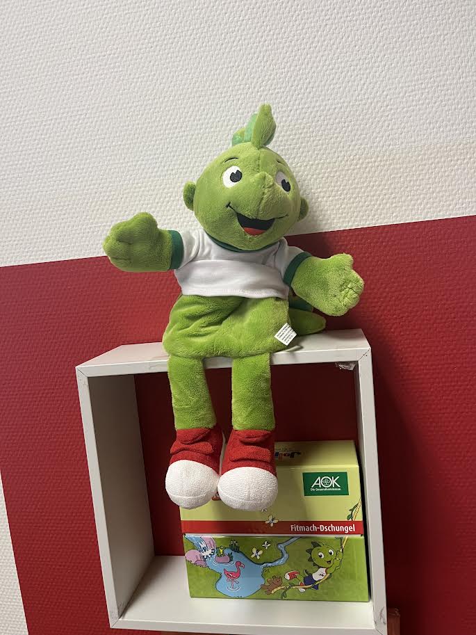 Auf dem Bild ist eine grüne Handpuppe mit freundlichem Gesicht zu sehen. Die Figur trägt ein weißes T-Shirt mit grünen Ärmeln sowie rote Schuhe und sitzt auf einem weißen Regal. Unter ihr steht eine bunte Box mit der Aufschrift „AOK – Fitmach-Dschungel“. Der Hintergrund ist eine zweifarbige Wand in Weiß und Rot.